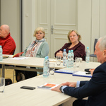 Grupa osób siedzi przy stole. Dwie kobiety patrzę w stronę kobiety i mężczyzny siedzących na szczycie stołu.