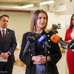 Kobieta w ciemnej marynarce mówi do mikrofonów. Za nią stoi kilka osób. 