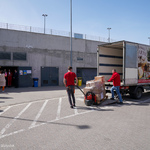 Parking, otwarta przestrzeń. Mężczyźni wypakowują paczki z ciężarówki. W tle budynki magazynowe. 