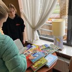 Dwie osoby stoją przy stole, na którym leżą kolorowe książki. Jedna z osób trzyma otwartą książkę. Światło wpada przez duże okno.  