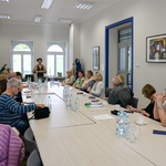 Jasne, przestronne pomieszczenie z dużymi oknami. Kilkanaście osób siedzi przy długim stole stojącym na środku pomieszczenia.