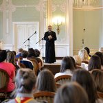 Wnętrze auli. Stojący na scenie ksiądz mówi do zgromadzonych..