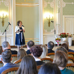 Wnętrze auli. Uczestnicy gali siedzą na krzesłach. Kobieta w jasnej marynarce mówi ze sceny do zebranych. 