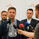 Mężczyzna w granatowej koszuli ozdobionej białym haftem mówi do kilku mikrofonów. Za nim stoi zastępca prezydenta i mężczyzna w okularach. 