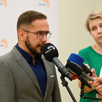 Mężczyzna w okularach mówi do kilku podstawionych mikrofonów.  