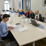 Jasne pomieszczenie. Przy stojącym na środku, długim stole siedzi kilkanaście osób. 