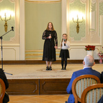 Dwie dziewczynki stoją na scenie trzymając w rękach mikrofony. Wyższa z dziewczynek trzyma za rękę młodszą. 