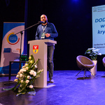 Scena Kina Forum. Prelegent mówi do zebranych stojąc przy mównicy. W tle, na ekranie, wyświetlona jest plansza prezentacji.