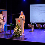 Scena Kina Forum. Dwie prelegentki stoją przy mównicy, jednak z nich mówi do mikrofonu. W tle, na ekranie, wyświetlona jest plansza prezentacji dotyczącej warsztatów dla rodziców..