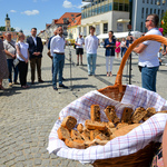 Na pierwszym planie kosz z kromkami chleba. W tle autor fotografii mówi do mikrofonu. Obok stoją inni uczestnicy wydarzenia. 