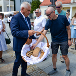 Prezydent miasta trzyma w dłoniach duży wiklinowy kosz z kromkami chleba, które rozdaje uczestnikom wydarzenia. Mężczyzna w okularach sięga po kawałek chleba. 