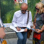 Autor fotografii składa autograf na broszurze pamiątkowej. Obok stoją uczestniczki wernisażu. 