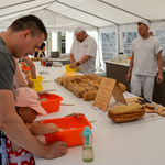 Namiot pod którym odbywają się warsztaty z pieczenia chleba. Długi stół na którym leżą chleby, noże i miski. Przy stole stoją uczestnicy warsztatów wygniatający ciasto oraz piekarze.  