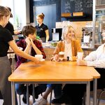 Wnętrze bistro: uczniowie siedzą przy stoliku. W tle uczennica pracuje za barem.