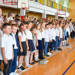 Uczniowie szkoły stoją w rzędzie na sali gimnastycznej podczas uroczystości rozpoczęcia roku.  