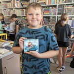 Dzieci w bibliotece przeglądają książki. Na pierwszym planie chłopiec trzyma audiobook 