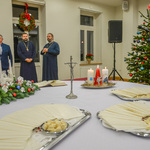 Duża, przestronna, jasna sala Centrum Aktywności Społecznej. Na pierwszym planie przykryty obrusem stół, na którym stoją tace z opłatkami, krzyż, świece i stroik. W tle stojący przy ścianie duchowni oraz udekorowana choinka.
